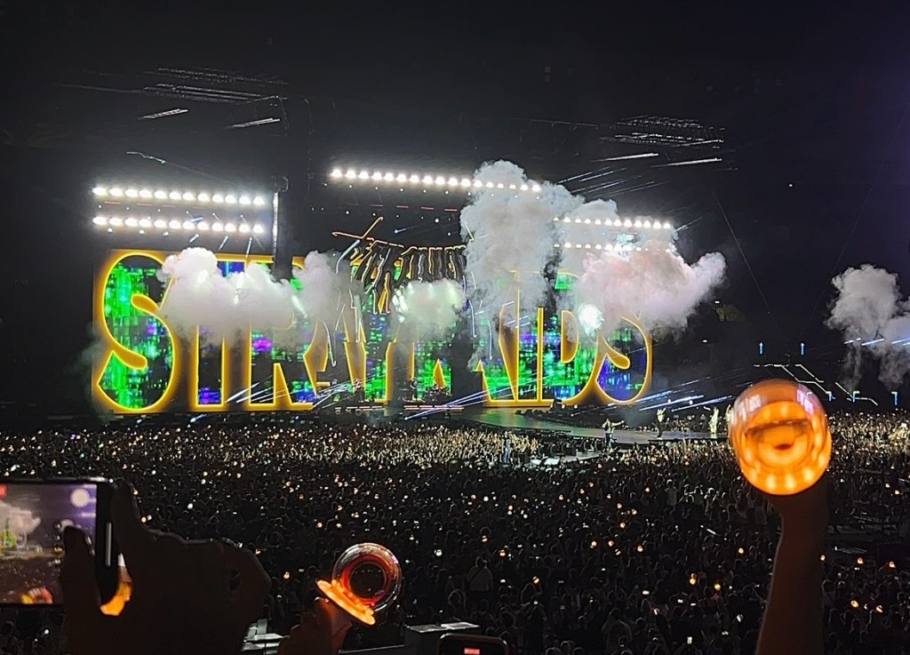Concerto degli Stray Kids allo Stadio Olimpico di Roma, con un pubblico di oltre 70.000 fan. Sul palco, il nome 'STRAY KIDS' è illuminato e circondato da effetti scenici e fumi, mentre i fan esprimono la loro energia con luce dei lightstick.